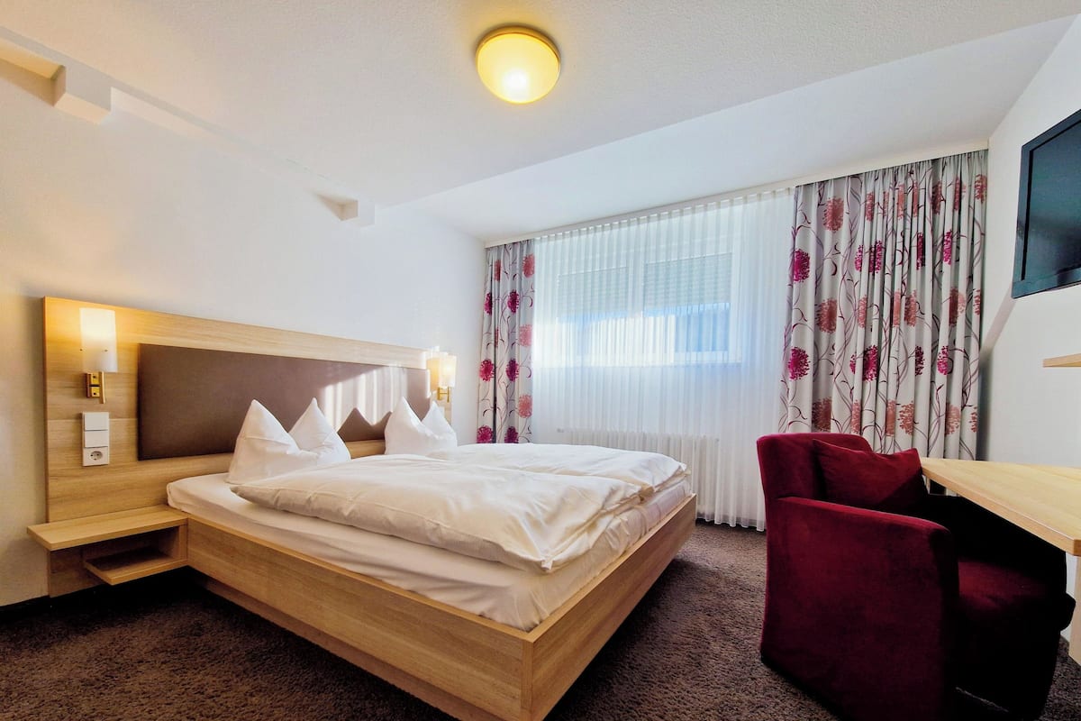 Une chambre d’hôtel avec un lit en bois, une chaise rouge, un bureau et une grande fenêtre ornée de rideaux à motifs floraux.