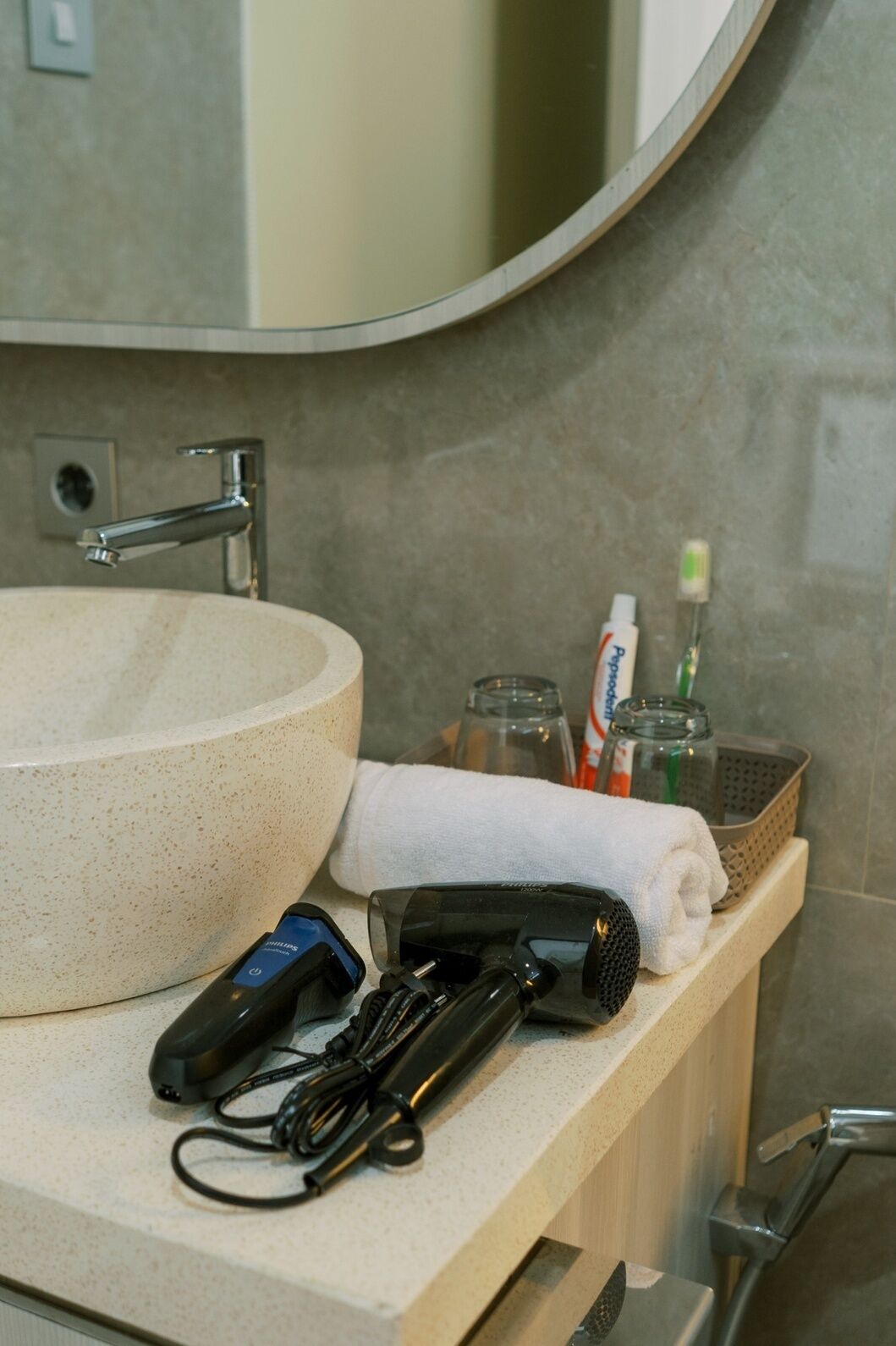 Un baño con un espejo redondo, un lavamanos, una toalla, un secador de pelo y un cepillo de dientes.