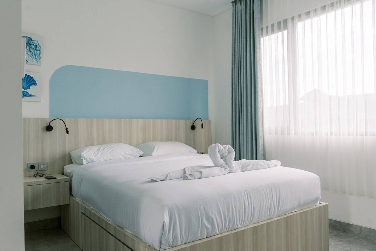 A modern hotel room with a large bed, a bedside table with a phone and charger, a wall-mounted lamp, and a window with curtains.