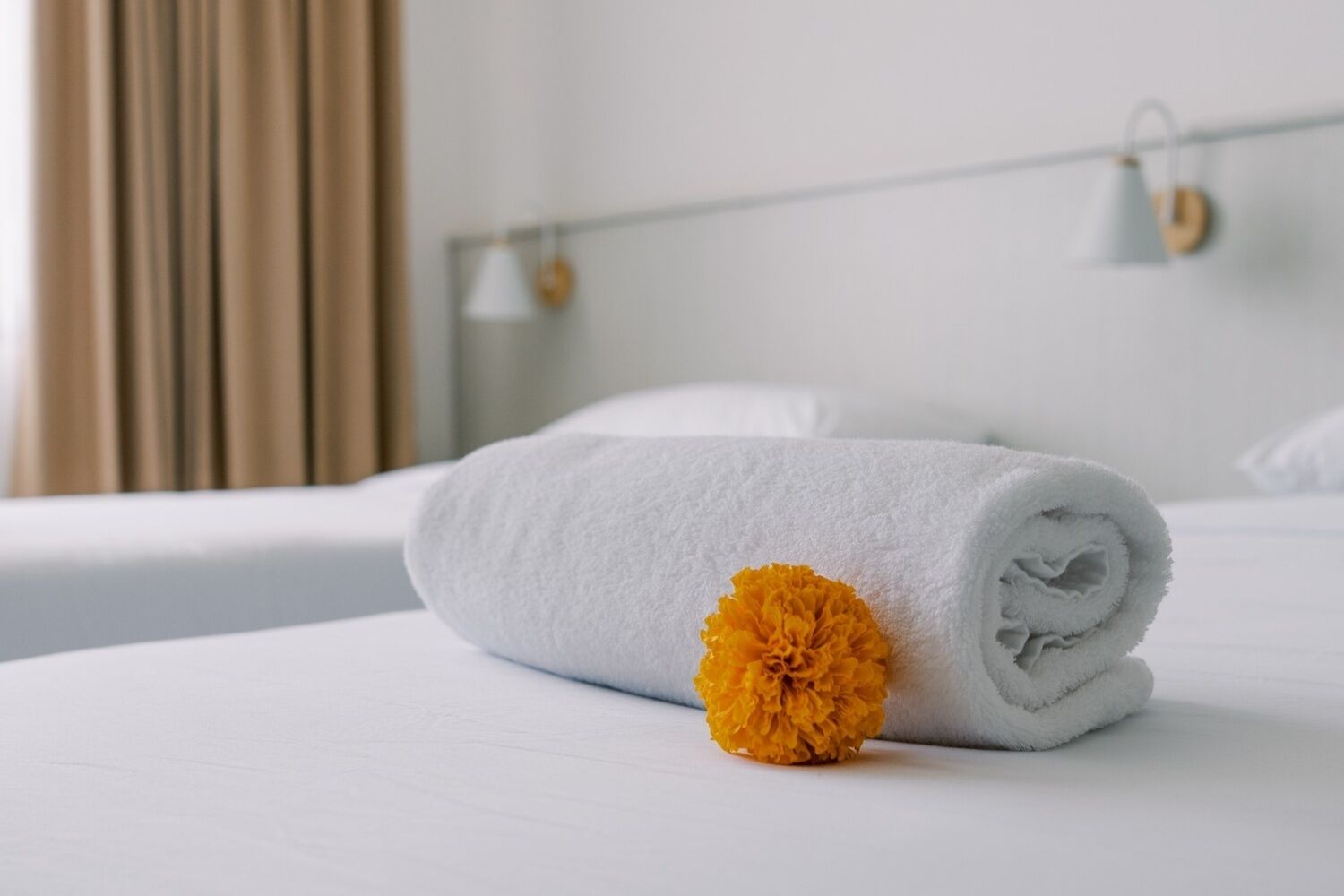 A neatly made bed with a rolled white towel and a single yellow flower on the bed.