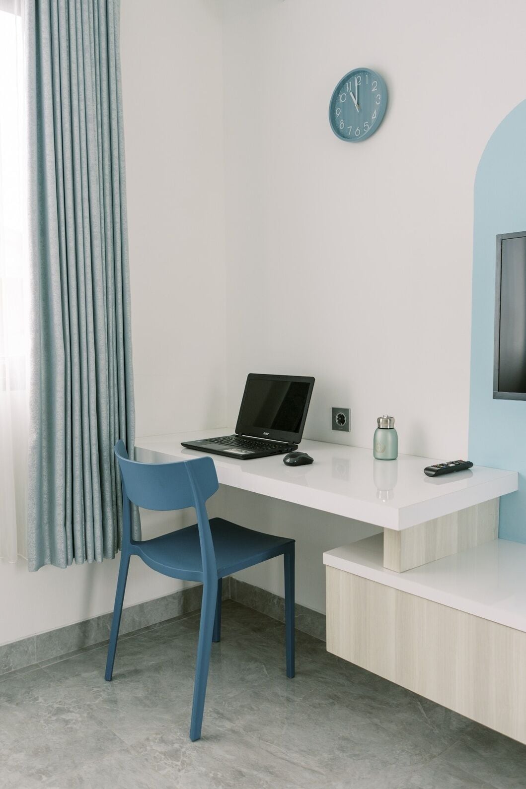 Una habitación moderna con un escritorio blanco, una silla azul, una computadora portátil, un reloj, un control remoto y un televisor.