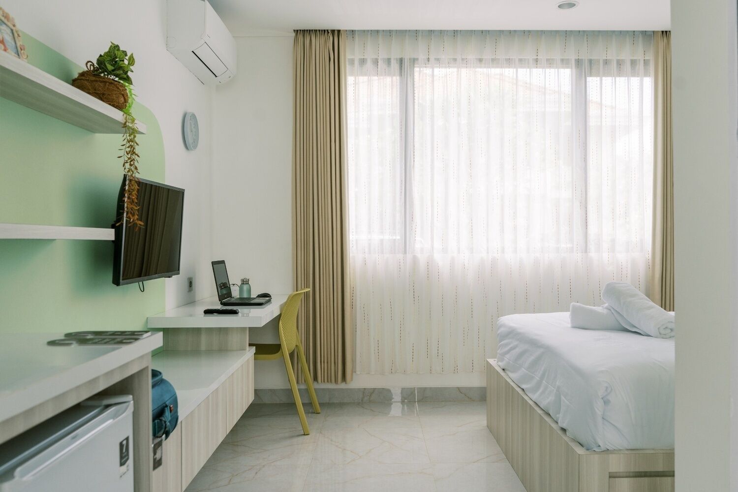 Une chambre d’hôtel avec un lit, un bureau équipé d’un ordinateur, une chaise, une télévision et une fenêtre avec des rideaux.