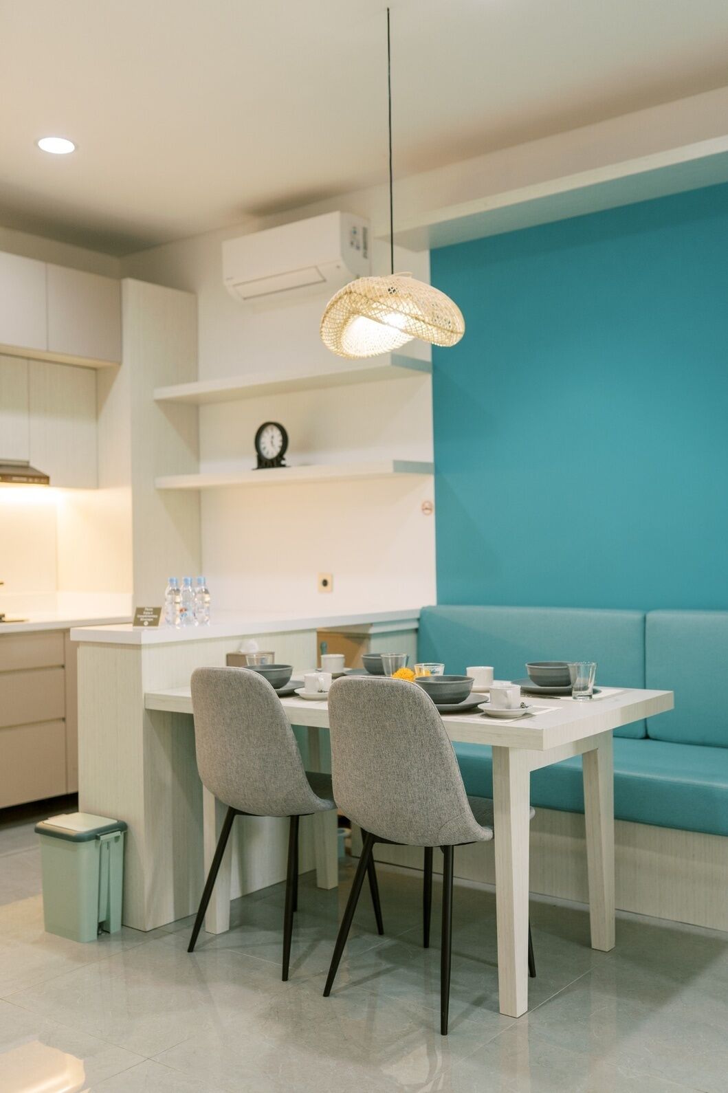 A modern kitchen with a dining area, featuring a teal accent wall, a hanging light fixture, and a small clock on the wall.