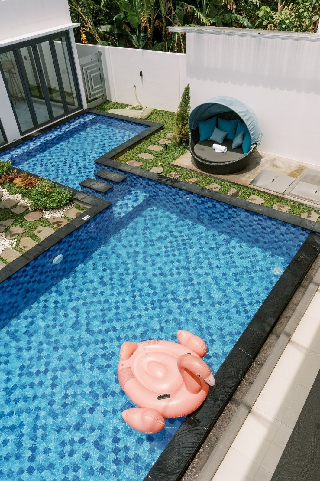 Una piscina con un flamenco inflable rosa, un espacio de descanso con cojines y una pequeña área ajardinada con piedras para pisar.