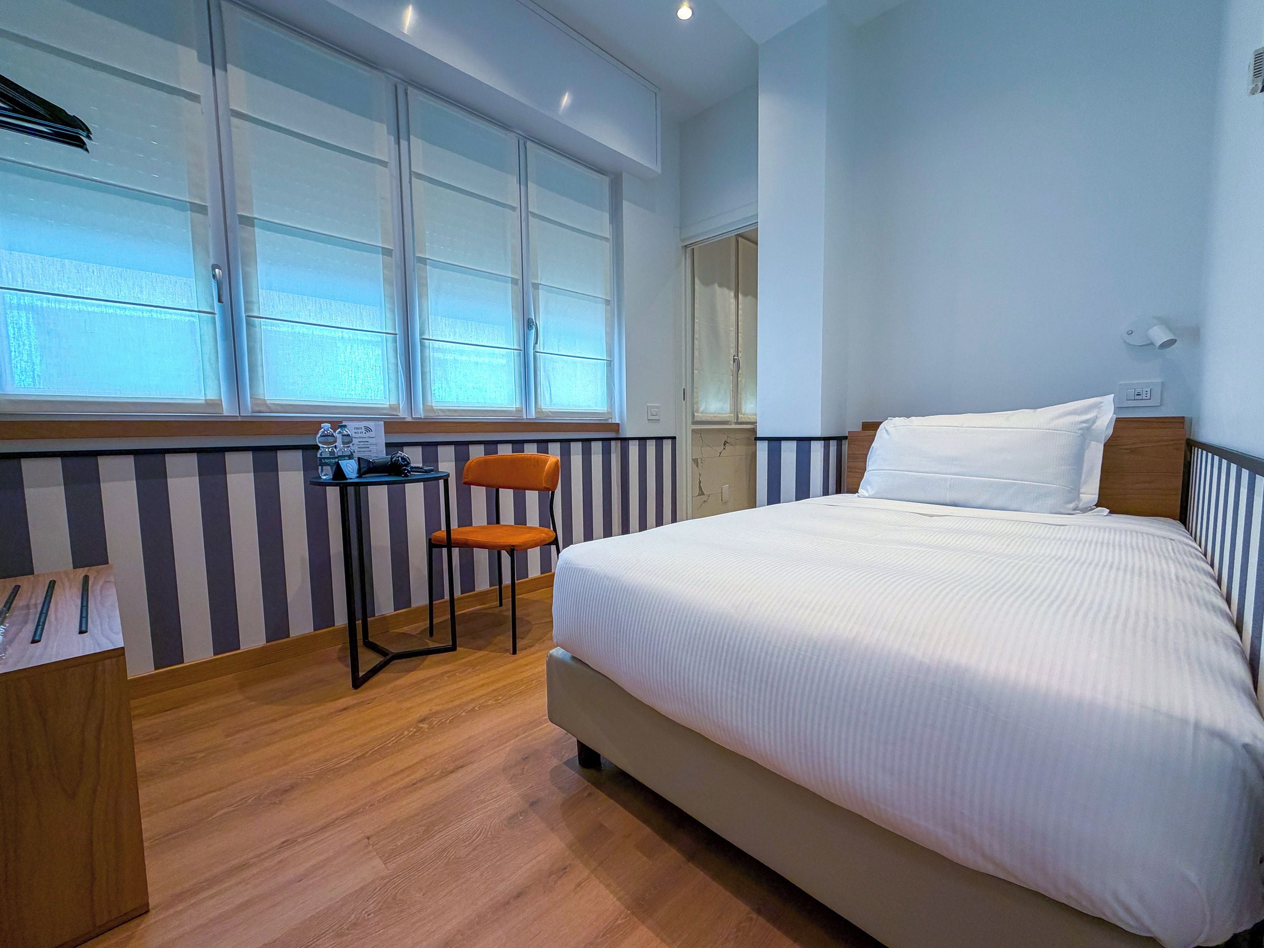 Une chambre d’hôtel avec un lit, une chaise, une petite table sur laquelle se trouvent une bouteille et des verres, et une fenêtre avec des rideaux.