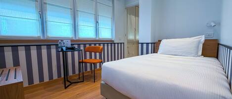 Et hotelværelse med seng, stol, et lille bord med flaske og glas samt et vindue med gardiner.