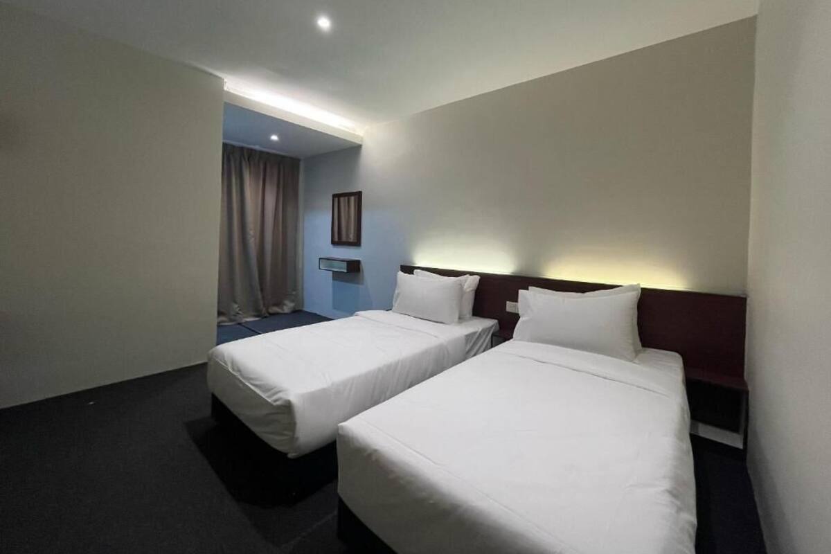 Une chambre d’hôtel avec deux lits simples, une moquette sombre, un mur de couleur claire et un luminaire au plafond.