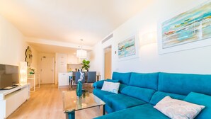 Ein modernes Wohnzimmer mit einem blauen Sofa, einem Glastisch und einem Fernseher auf einem weißen Lowboard. Im Hintergrund ist eine Küchenzeile mit weißen Schränken und einem Esstisch zu sehen.
