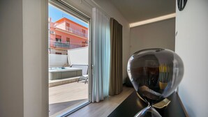 A modern hotel room with a sliding glass door leading to a balcony with a hot tub and a view of a red building. - Mediterranea Suite (Avola)