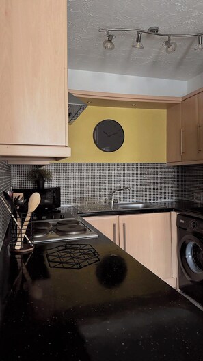 A kitchen with a black countertop, a sink, a stove, a washing machine, and a clock on the wall.