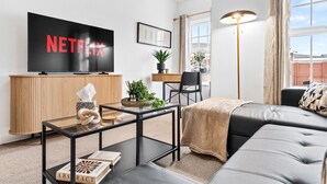 A modern living room with a flat-screen TV displaying 'NETFLIX', a sofa, a coffee table, a side table with a plant, and a floor lamp. - Modern 2-Bed Flat with Flexible Beds (Warrington)