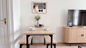 A dining area with a wooden table, benches, a vase with flowers, and a TV on a cabinet. - Modern 2-Bed Flat with Flexible Beds (Warrington)