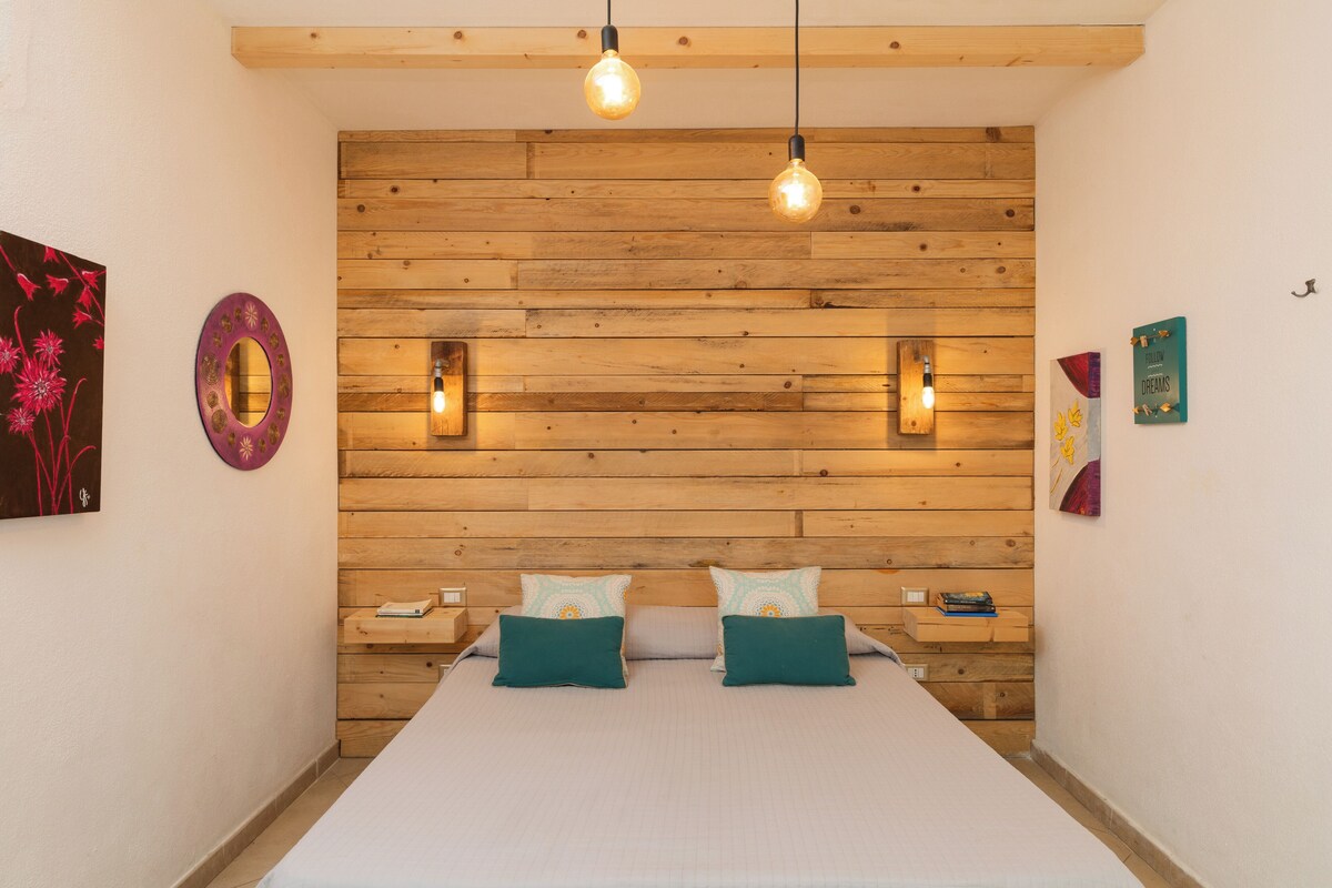 Een slaapkamer met een houten hoofdbord, twee nachtkastjes, een bed met teal kussens en wandlampen.