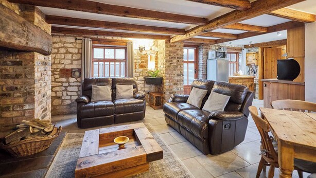 Ein rustikales Wohnzimmer mit Steinwänden, Holzbalken, einem Ledersofa, einem Holztisch und einem Essbereich mit Holztisch und Stühlen.