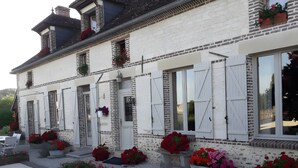 Un edificio blanco con detalles de ladrillo, numerosas ventanas con contraventanas y jardineras con flores a lo largo de la fachada.