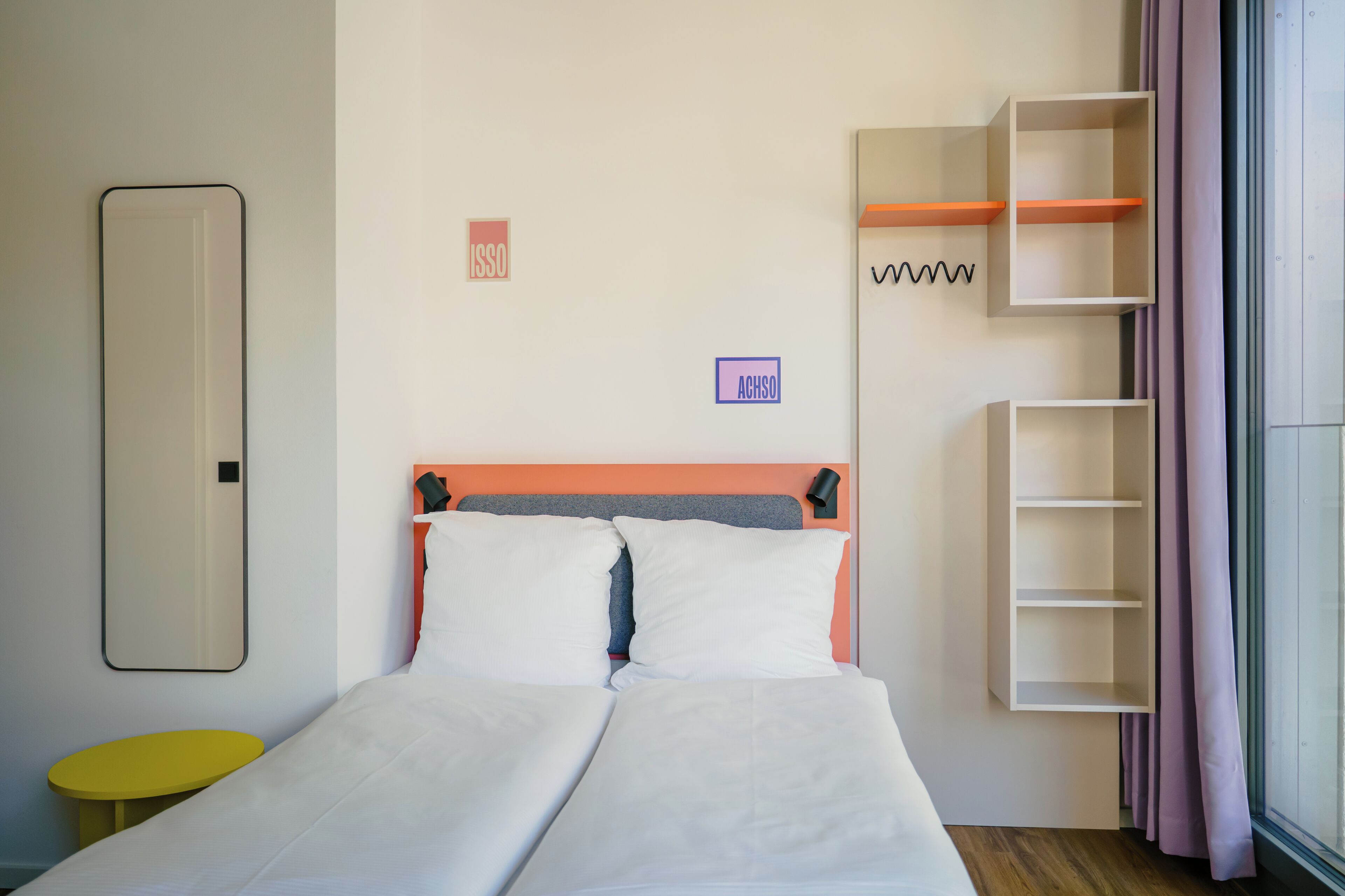 Ein Doppelbett mit weißer Bettwäsche, ein kleiner gelber Nachttisch und eine an der Wand montierte Regalunit mit einer Lampe.