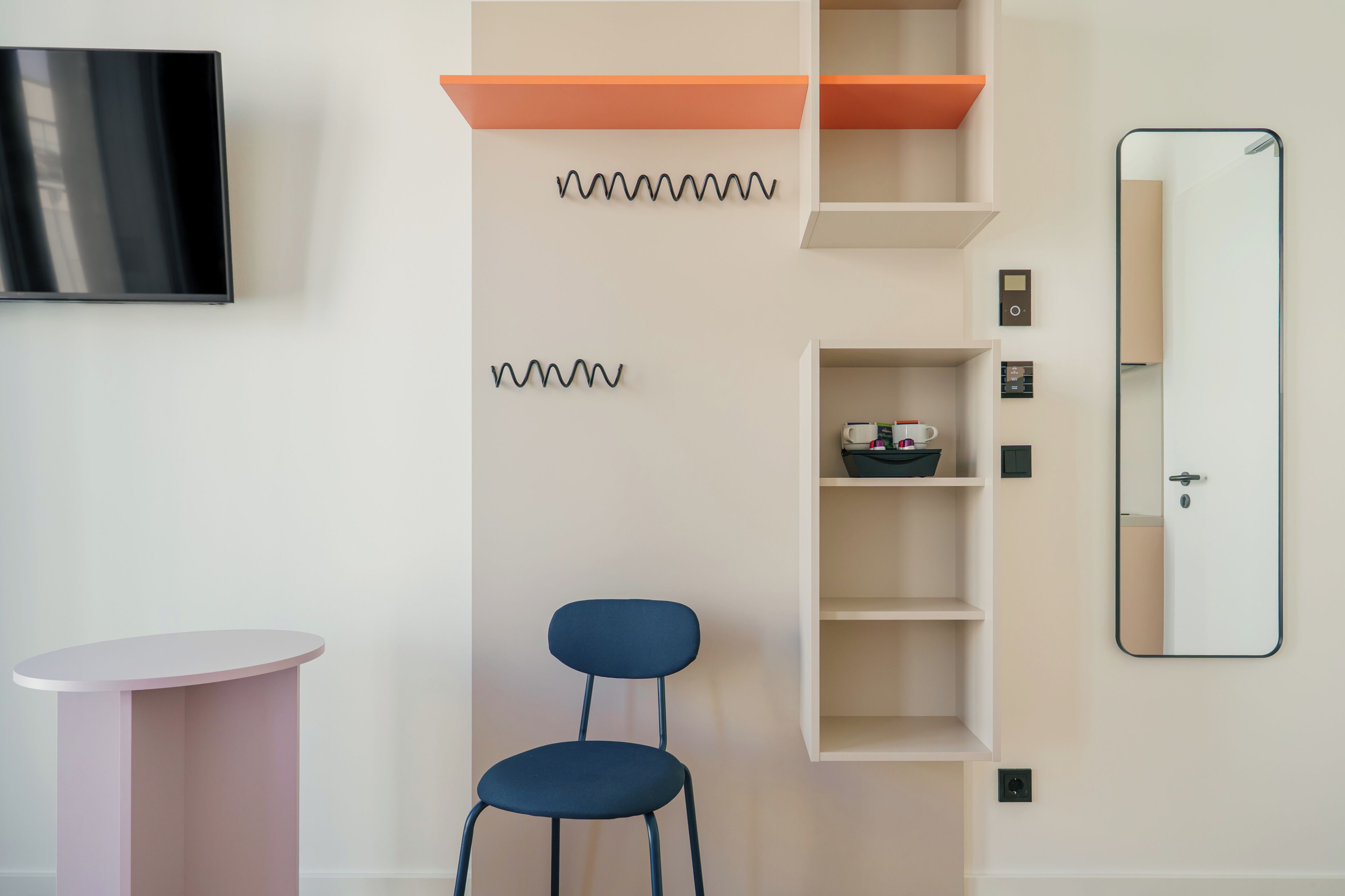 Ein modernes Zimmer mit Flachbildfernseher, einem runden rosafarbenen Tisch, einem blauen Stuhl, einer weißen Regaleinheit mit orangefarbenen Akzenten und einem Spiegel.