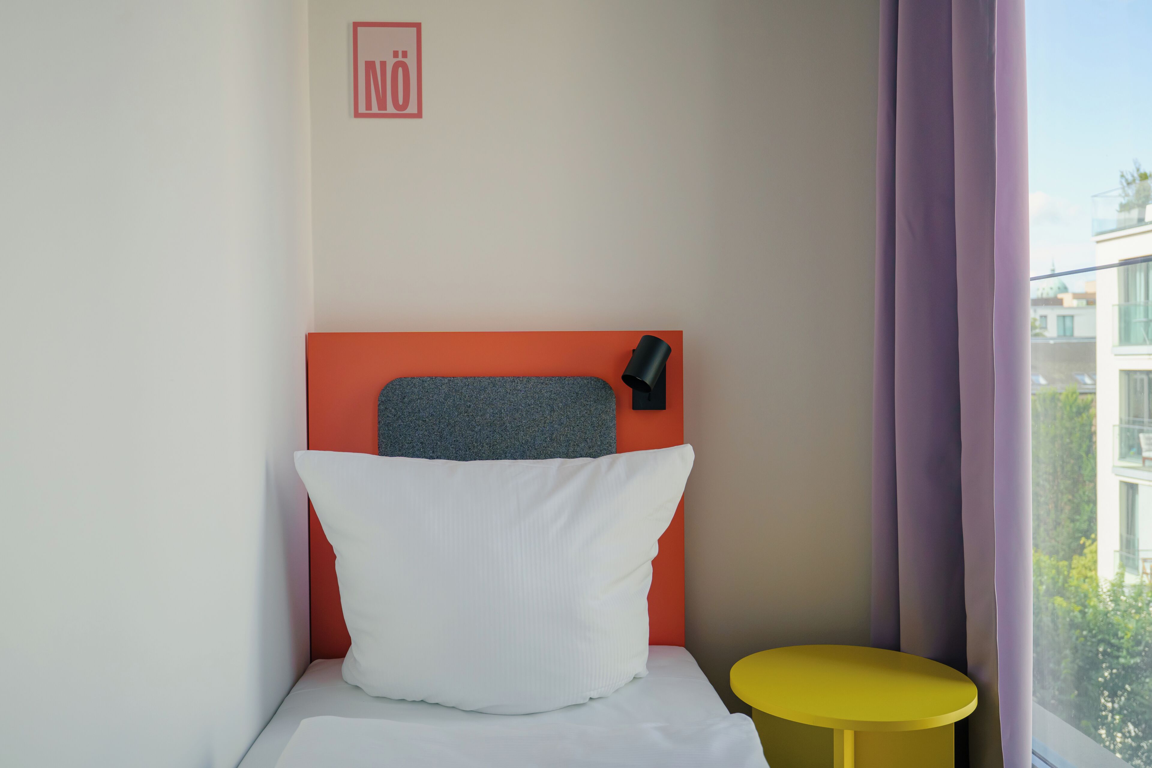 A hotel room with a bed, a small round yellow table, a window with purple curtains, and a city view.