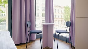 A room with two chairs and a small table, a window with purple curtains, and a view of buildings outside. - Homaris K48 Studios (Berlin)