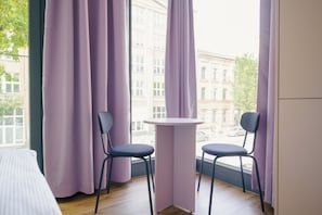 A room with two chairs and a small table, a window with purple curtains, and a view of buildings outside. - Homaris K48 Studios (Berlin)