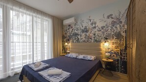 A bedroom with a large bed, a nightstand, a small table with a lamp, and a wall-mounted air conditioner. - Welcoming 1-bedroom apartment  in charming Sozopol (Sozopol)