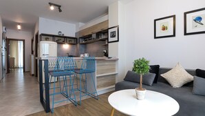 A modern kitchen with a built-in fridge, wooden cabinets, and a dining area with blue chairs. There is a round white table with a potted plant and a grey sofa with cushions. - Welcoming 1-bedroom apartment  in charming Sozopol (Sozopol)