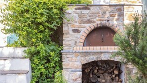 A stone-built outdoor pizza oven with a brick arch, surrounded by greenery and a stack of firewood.