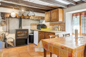 A kitchen with a wood stove, wooden cabinets, and a dining table with a fruit-patterned tablecloth. - Small House on 5 Hectares with Pool, Sauna, Gym, Horses, Donkey (Osse-en-Aspe)