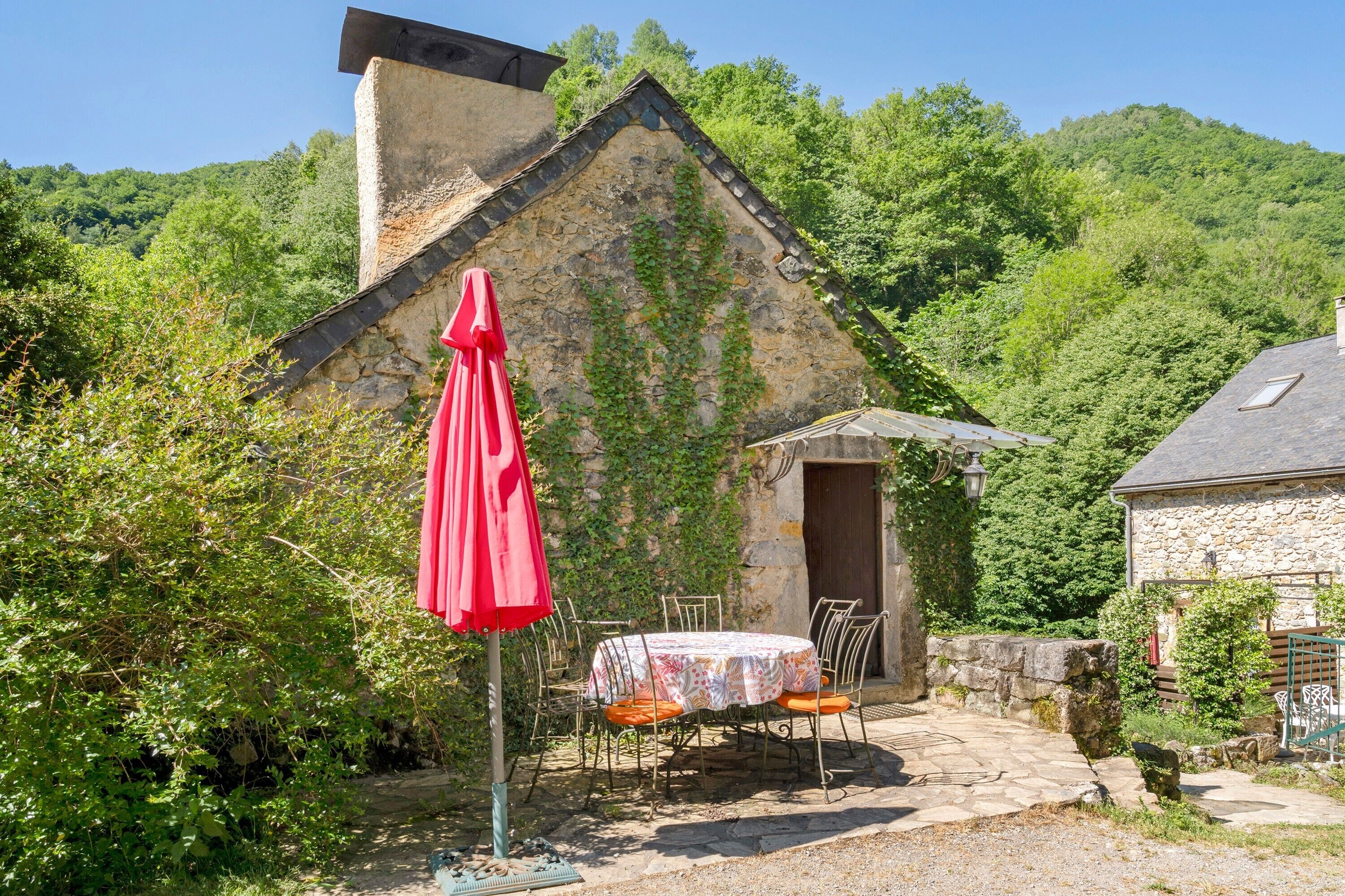 Um edifício de pedra com telhado inclinado, uma mesa com cadeiras e um guarda-sol vermelho.