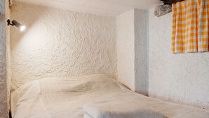 Une pièce avec un mur blanc à texture, un lit recouvert d’une couverture blanche, un élément décoratif en pierre et un rideau à carreaux.