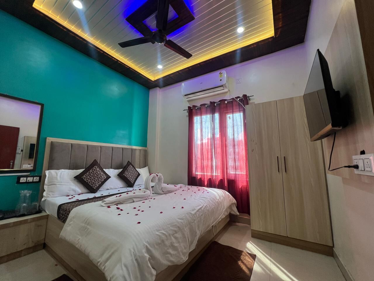 A modern hotel room with a bed, a ceiling fan, an air conditioning unit, a TV mounted on the wall, and a window with red curtains.
