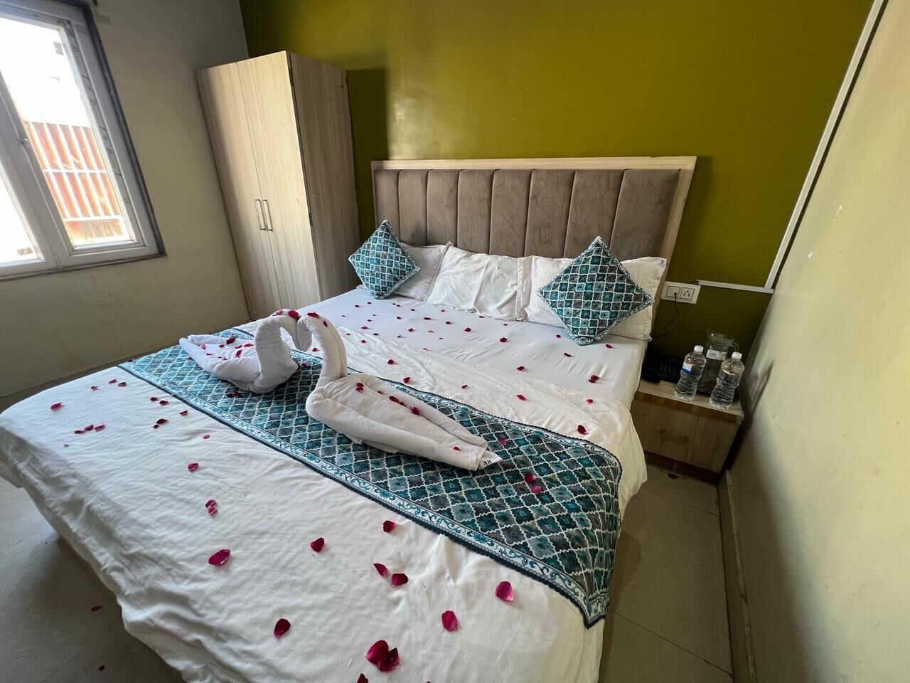 A neatly made bed with a swan-shaped towel and rose petals, a wooden headboard, and a bedside table with water bottles.