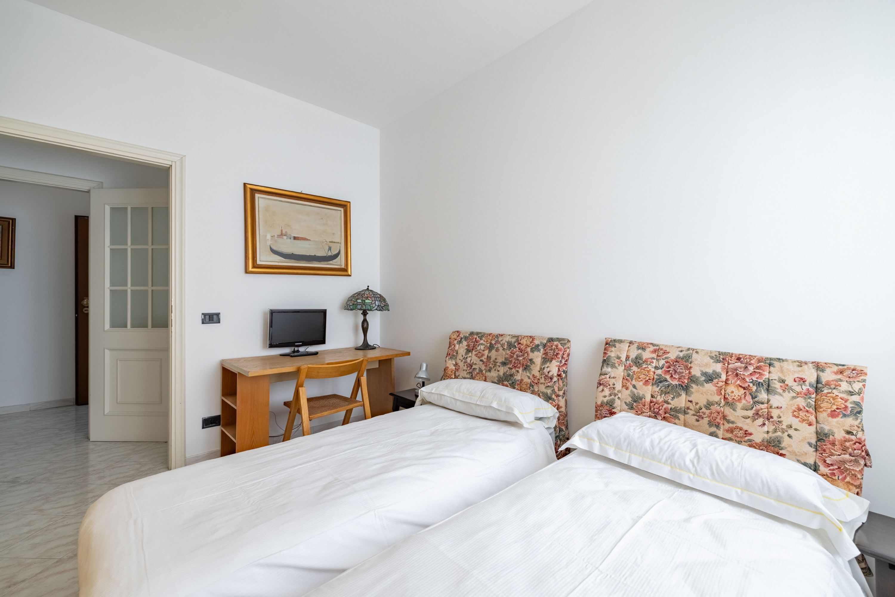 A bedroom with two beds, a desk with a computer, a lamp, and a painting on the wall.
