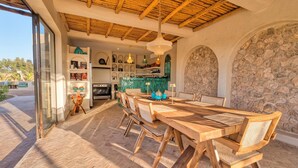 A dining area with a long wooden table and chairs, a kitchen in the background, and a stone wall. - La villa DUCASSE (Ounagha)
