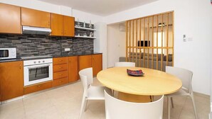 Uma cozinha moderna com armários de madeira, forno embutido e micro-ondas. Há uma área de refeições com uma mesa redonda e cadeiras brancas.