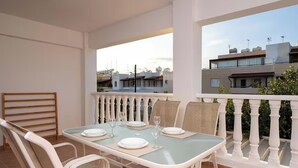 Varanda com mesa posta para refeições, cadeiras brancas e vista para prédios ao fundo.