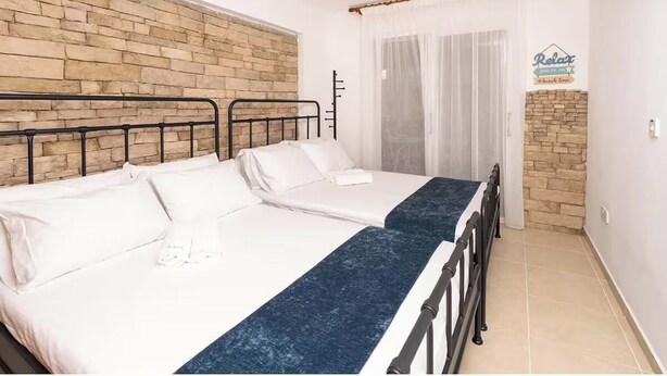 Quarto duplo com duas camas, parede de destaque em pedra, um letreiro com a palavra 'Relax' e uma janela com cortinas translúcidas.