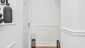 Un couloir avec une porte blanche, un appareil fixé au mur, un grand vase et une poubelle.