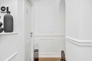 Un pasillo con una puerta blanca, un dispositivo en la pared, un jarrón grande y un cubo de basura.
