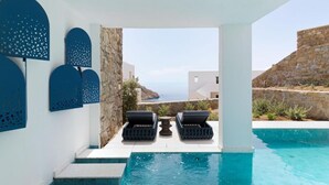Zona de piscina con escalones que bajan al agua, dos sillones de descanso y vista al mar y edificios al fondo.