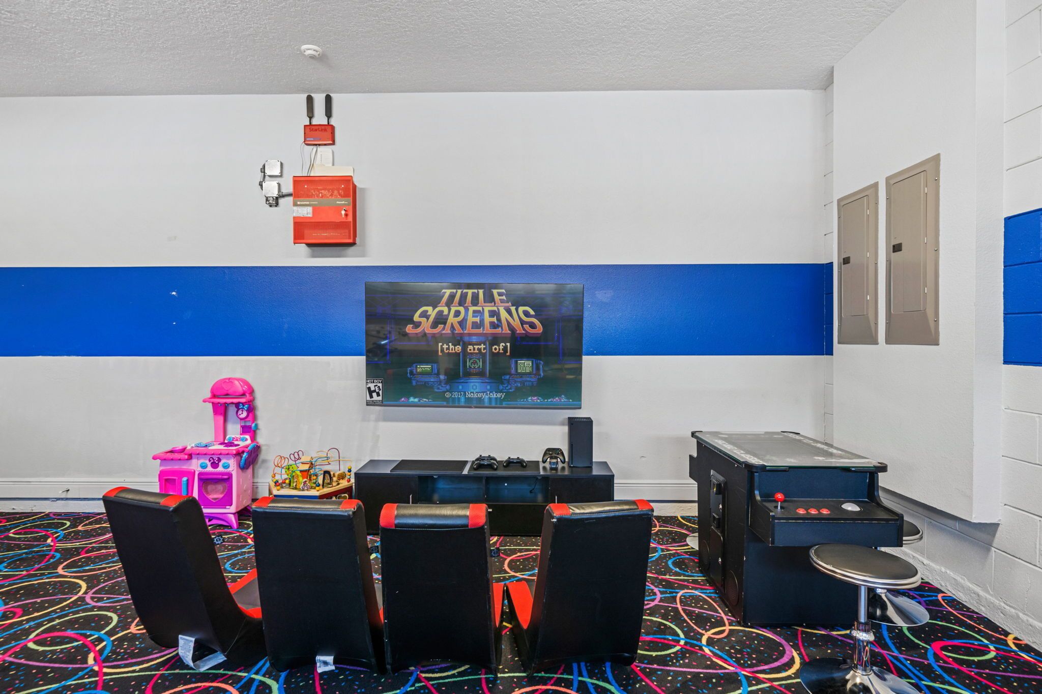 A room with a large screen displaying 'TITLE SCARES', gaming chairs, a pink claw machine, and a black arcade machine.