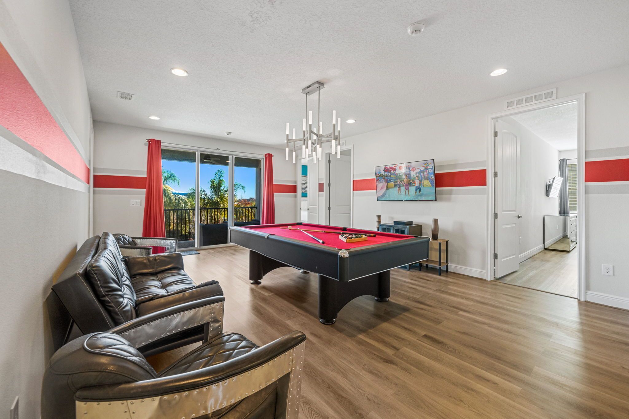 A pool table in a room with a wooden floor, a leather couch, and a door leading to a balcony with a view of palm trees.
