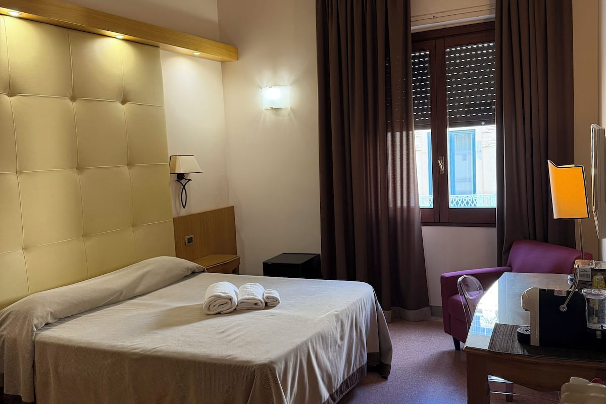 Habitación de hotel con una cama grande, un escritorio con silla, una lámpara y una ventana con cortinas.