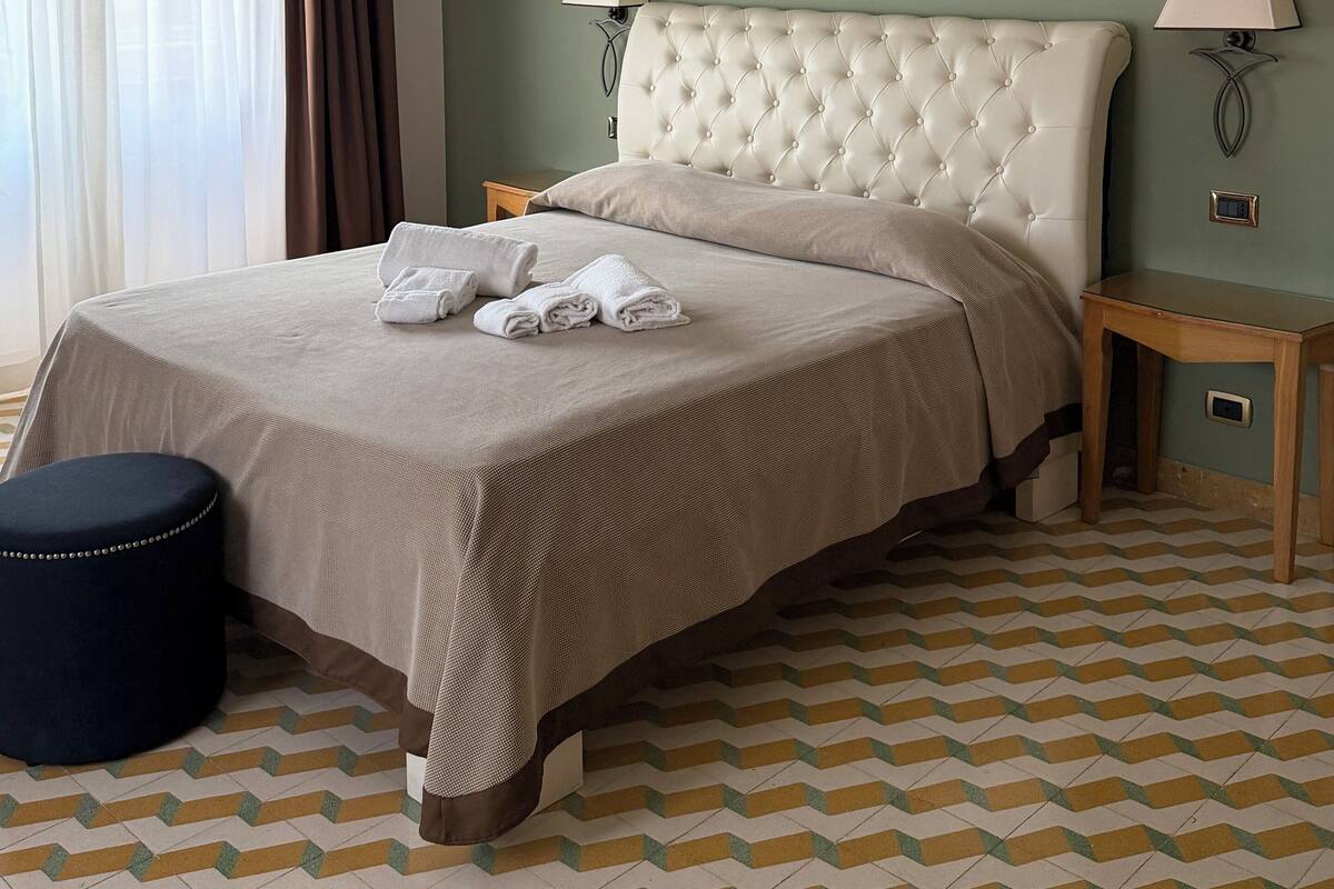 Una cama bien tendida con un cabecero capitonado, dos lámparas de noche y un par de toallas dobladas sobre la cama.