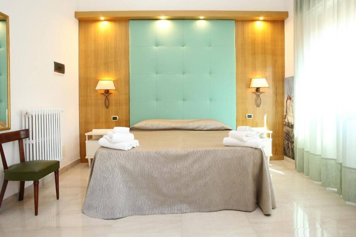 Habitación de hotel con una cama grande, dos lámparas de noche, un cabecero de madera, una silla verde y un radiador.