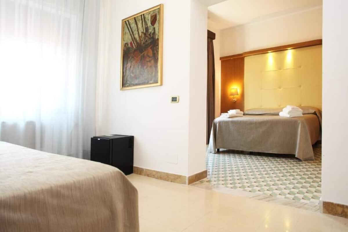 Habitación de hotel con cama, un cuadro en la pared, una mesita de noche con lámpara y suelo de baldosas.