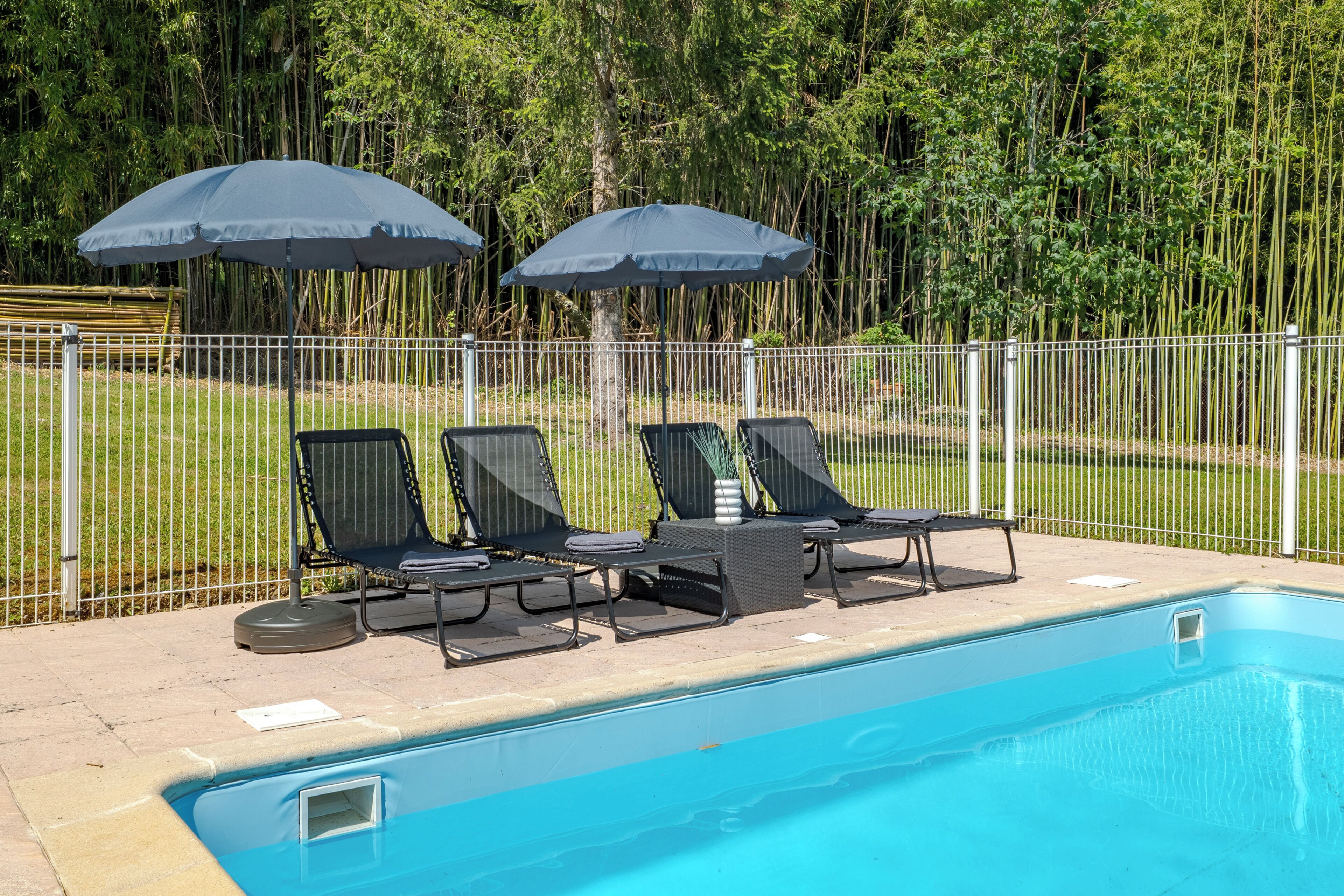 Une piscine entourée d’une clôture blanche et de verdure, avec des chaises longues et des parasols.