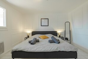 Un lit bien fait, recouvert d’une couvre-lit à motifs, avec deux serviettes enroulées et un seul oreiller. On y trouve des lampes de chevet, un certificat encadré accroché au mur et un grand miroir.