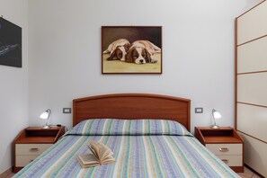 Une chambre à coucher avec une tête de lit en bois, une couvre-lit à motifs, deux tables de chevet avec des lampes et un tableau représentant des chiens accroché au mur.
