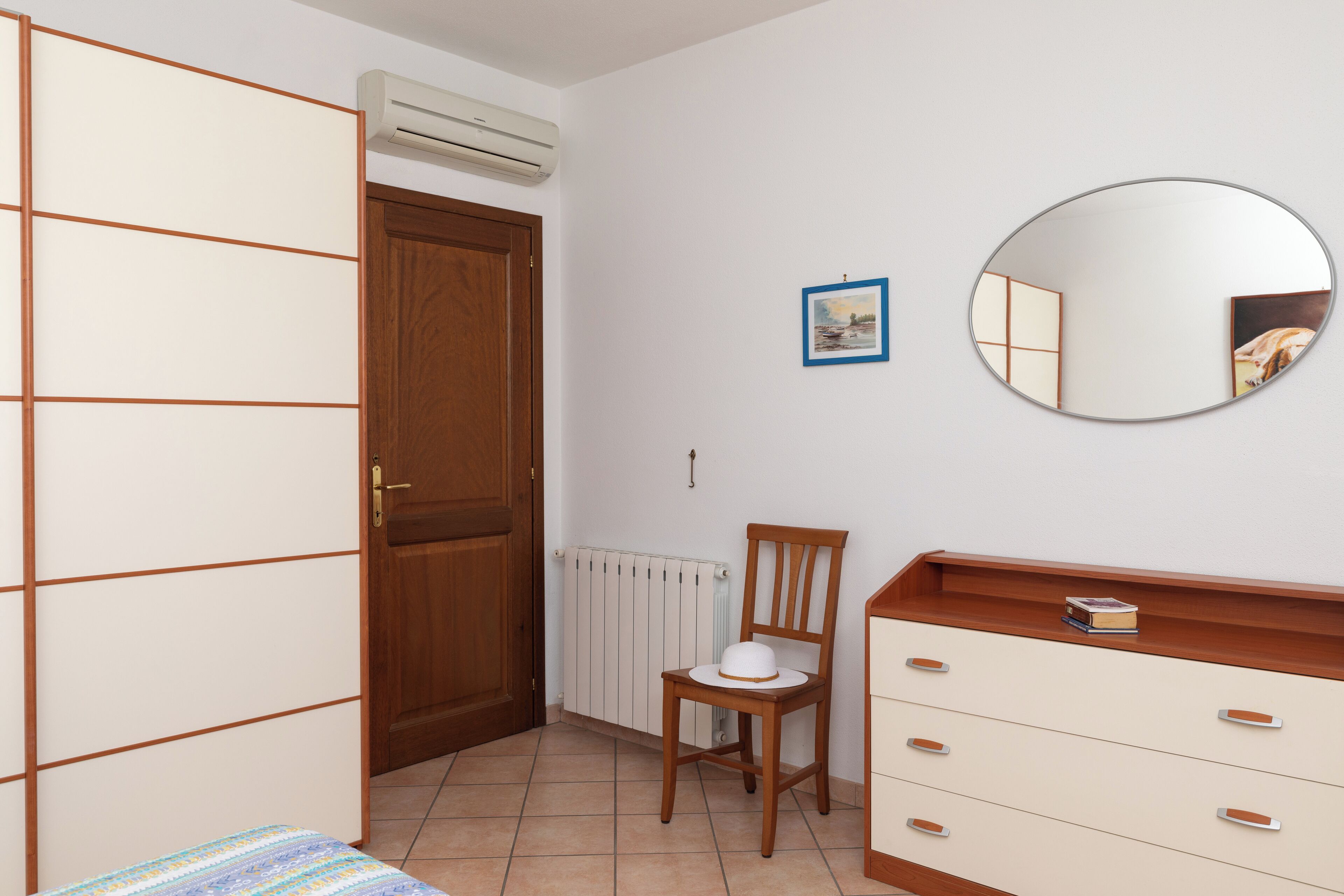 A bedroom with a wooden wardrobe, a chair, a dresser, a mirror, and a radiator.